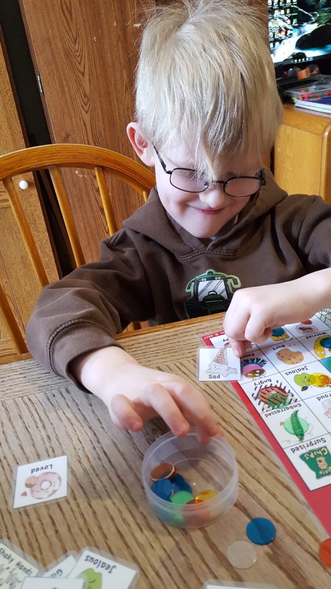 young blond boy in glasses holding a card to match on his bingo sheet reaching for a place marker haley sez