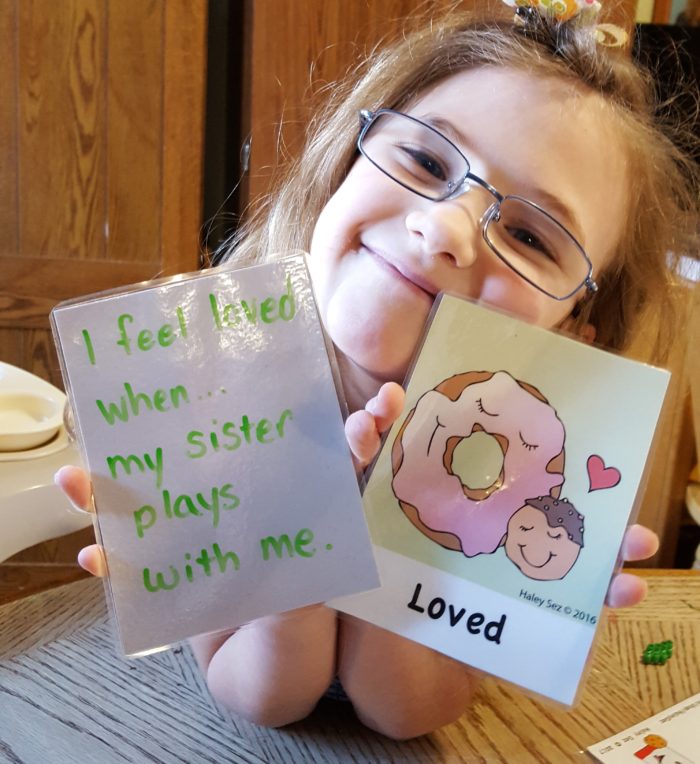 girl wearing glasses holding feeling card Loved and card that says I feel loved when my sister plays with me from haley sez