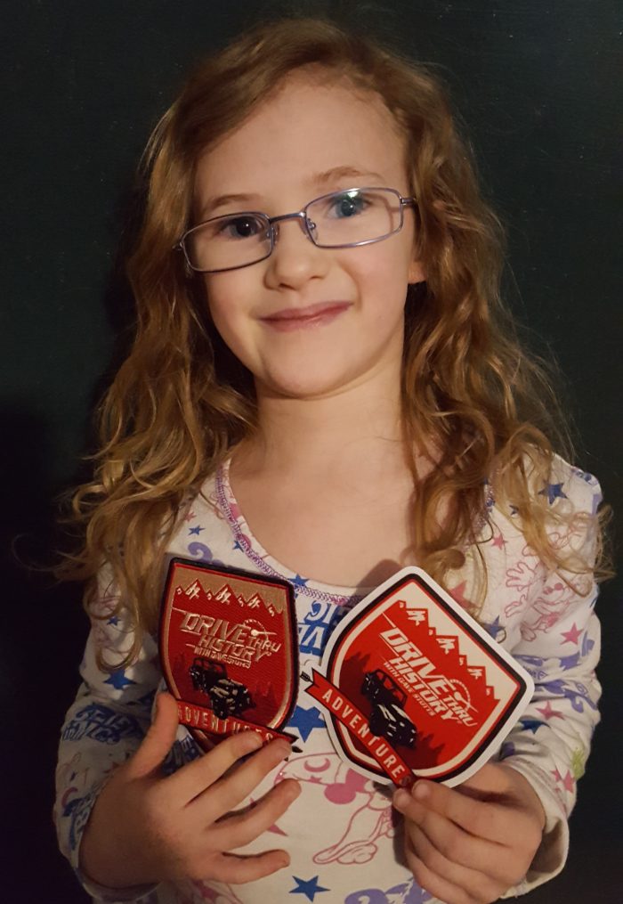 a young girl with curly hair and glasses holds a patch and sticker from Drive Thru History Adventures