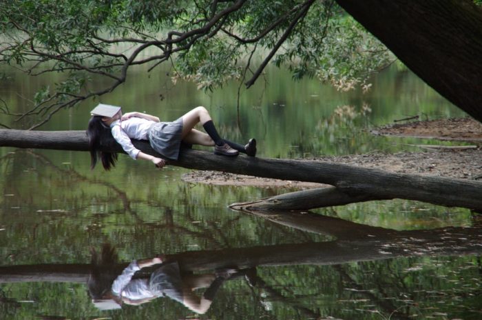 woman laying on a log with book on face above a lake trying something new for your down time