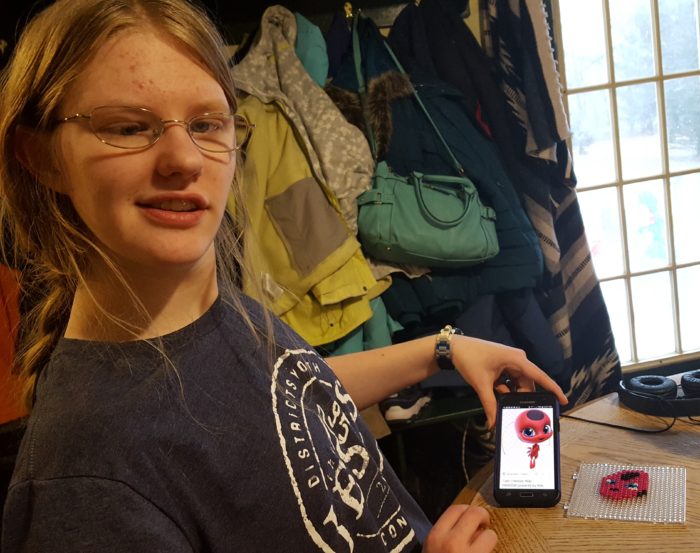 A blond adult woman with glasses with her smartphone photo of Miraculous Tikki and design board with Zirrly Super Beads product review