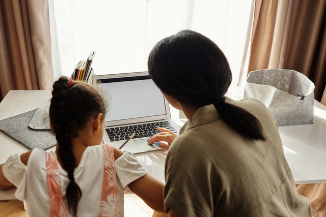 photo looking behind a mother and daughter doing homeschool with a laptop at a desk in front of an open window with curtains eliminating boredom