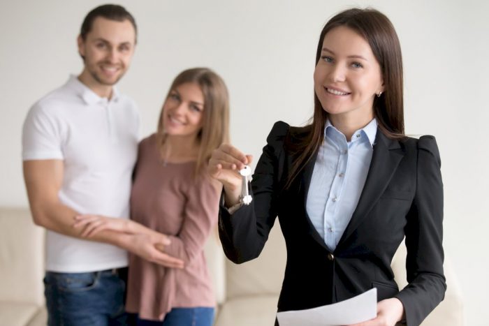 landlord holding key to rental property with smiling couple looking on from behind