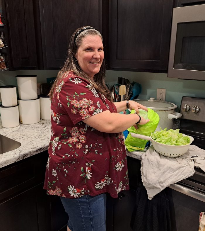 smiling woman in a kitchen opening the saladzilla salad spinner with lettuce in a colander next to it