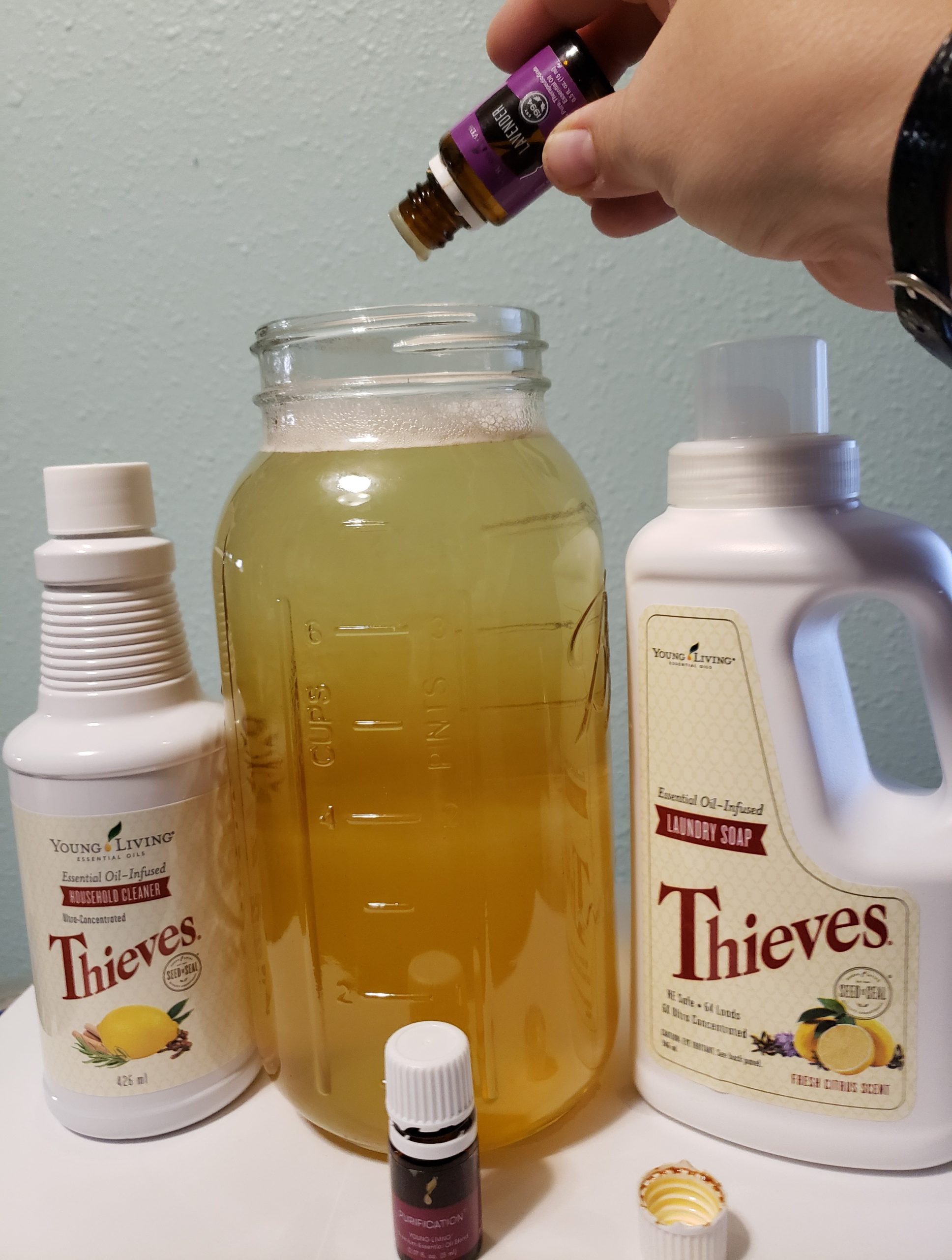 young living lavender essential oil being carefully dripped into a large glass jar of amber colored fluid next to thieves household cleaner laundry soap purfication blend
