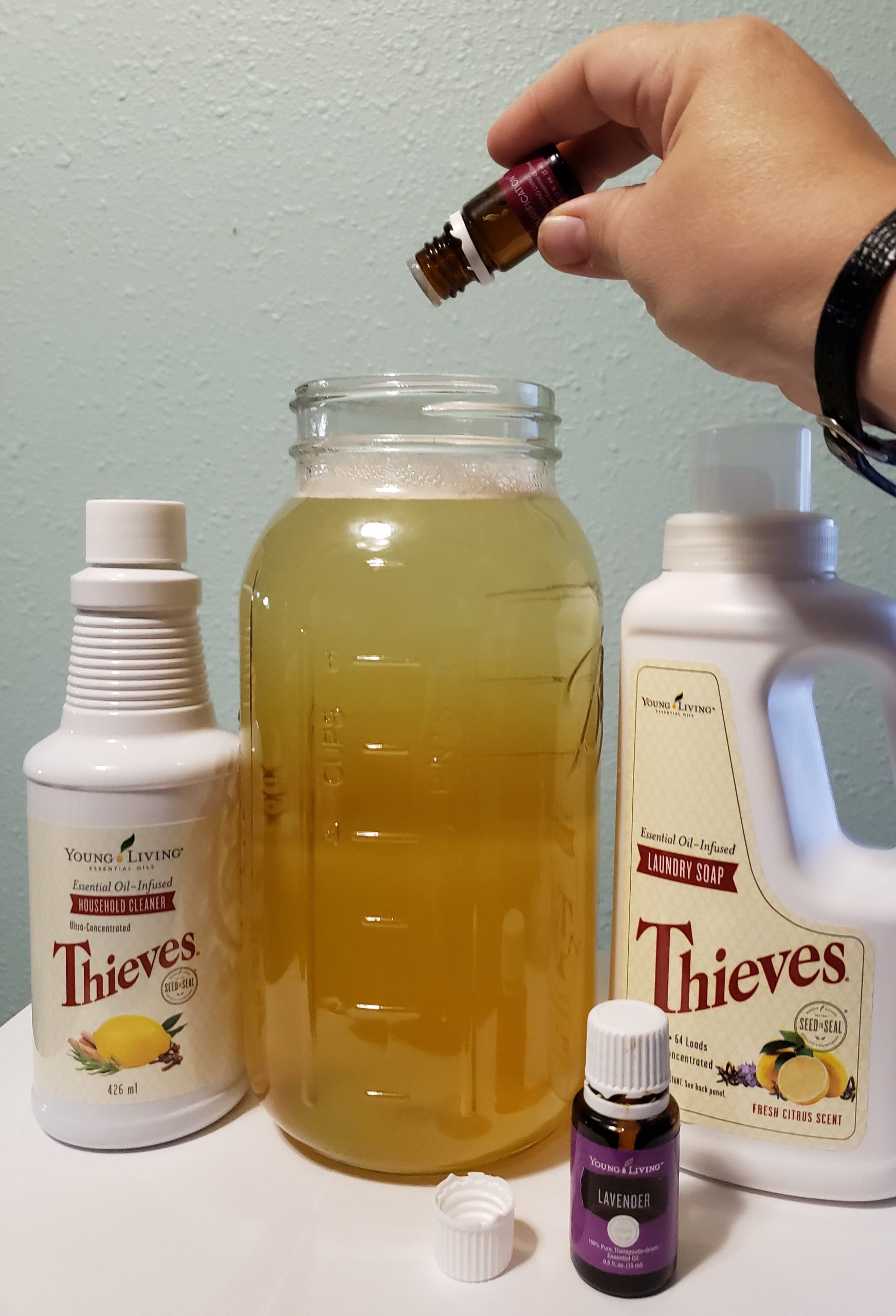 young living purification oil blend being carefully dripped into a large glass jar of amber colored fluid next to thieves household cleaner laundry soap lavender essential oil
