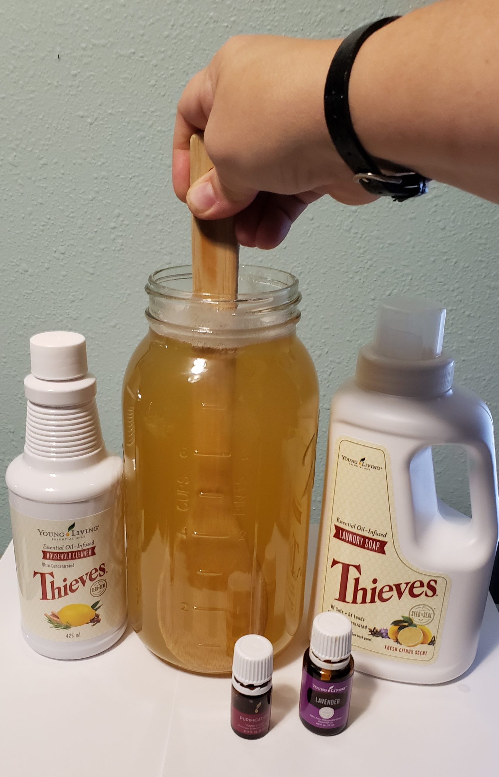 hand stirring wooden spoon to mix amber fluid contents inside a large glass jar next to young living thieves laundry soap thieves household cleaner and purification blend and lavender essential oil