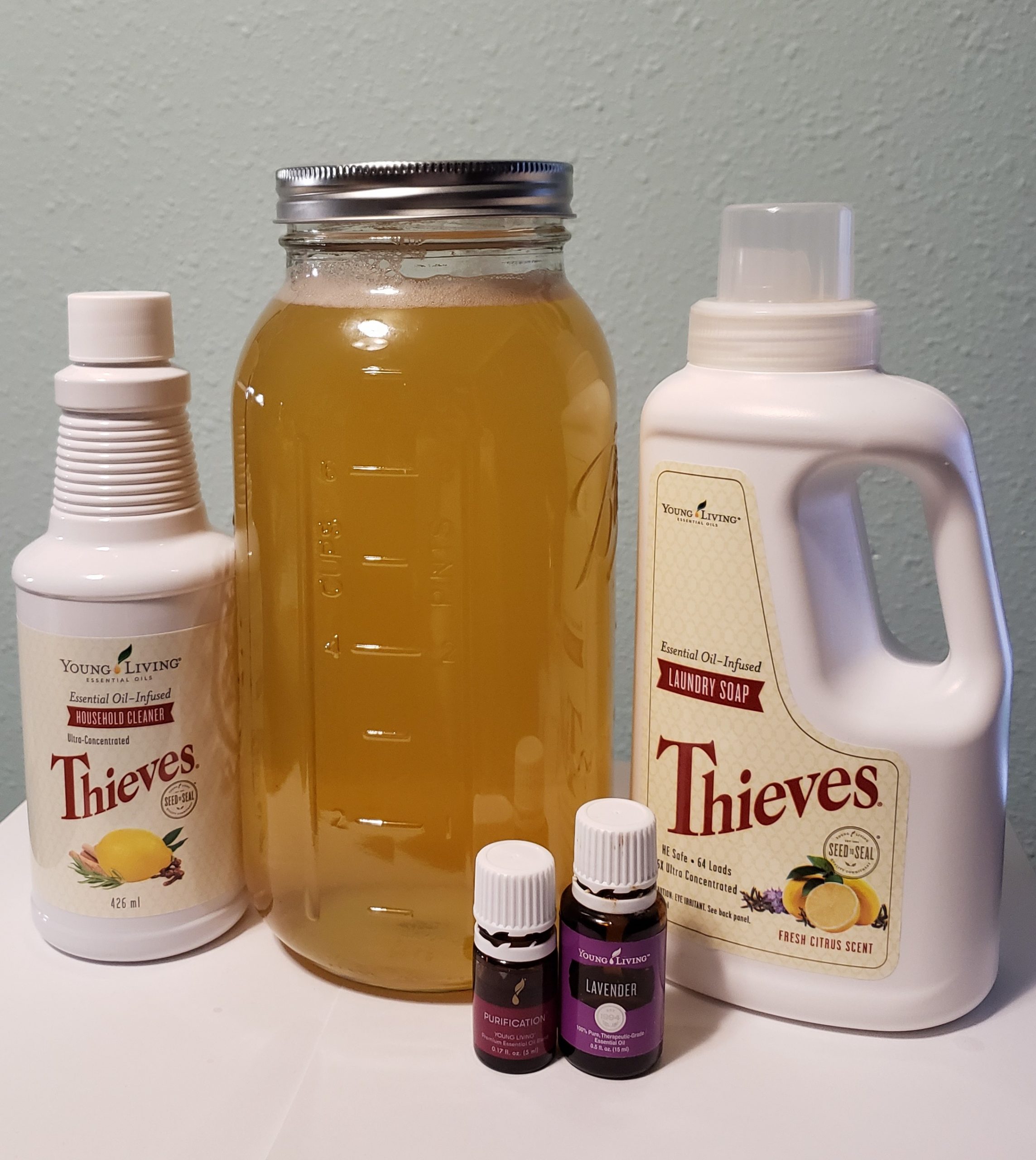 young living thieves household cleaner next to glass jar filled with amber fluid and lid on next to thieves laundry soap and purification blend and lavender essential oil