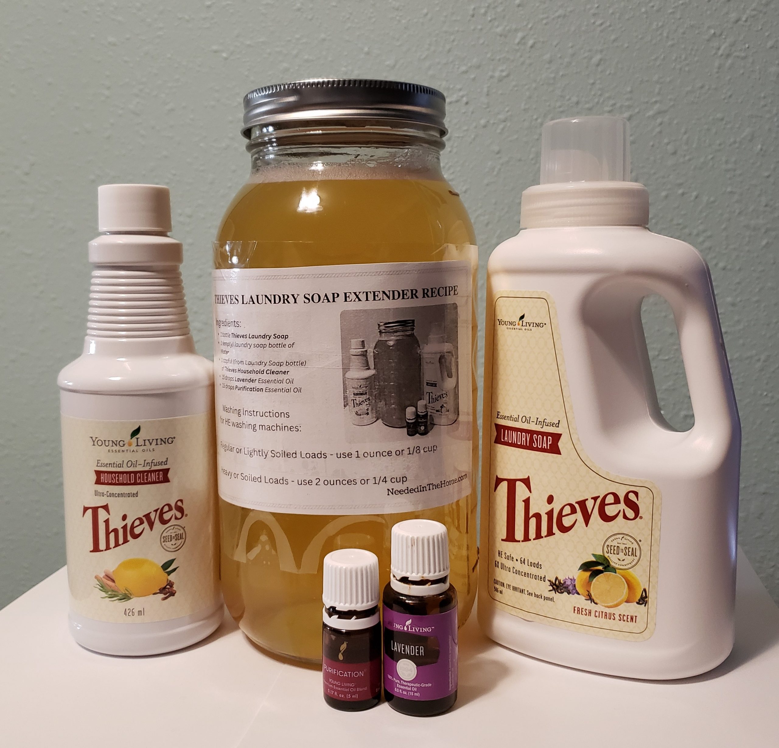 young living thieves household cleaner next to glass jar filled with amber fluid with recipe taped to front and lid on next to thieves laundry soap and purification blend and lavender essential oil
