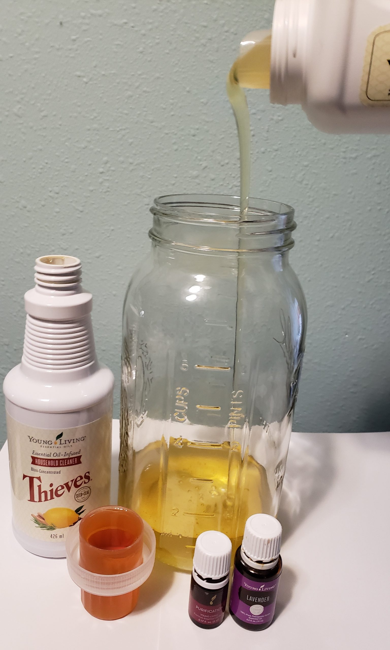 young living thieves laundry soap being poured into large glass jar next to thieves household cleaner and purification blend and lavender essential oil