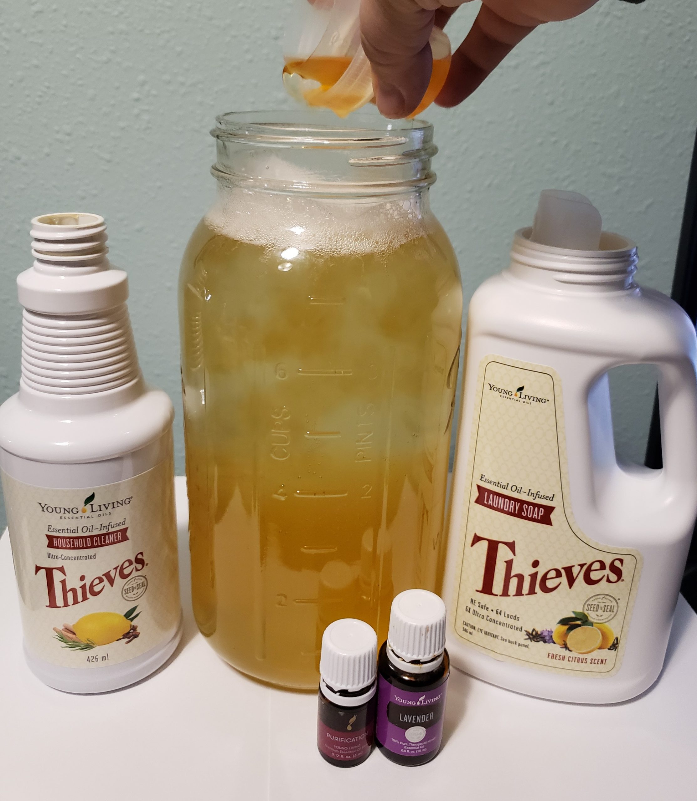 young living thieves household cleaner being poured into large glass jar next to thieves laundry soap and cleaner bottles and purification blend and lavender essential oil