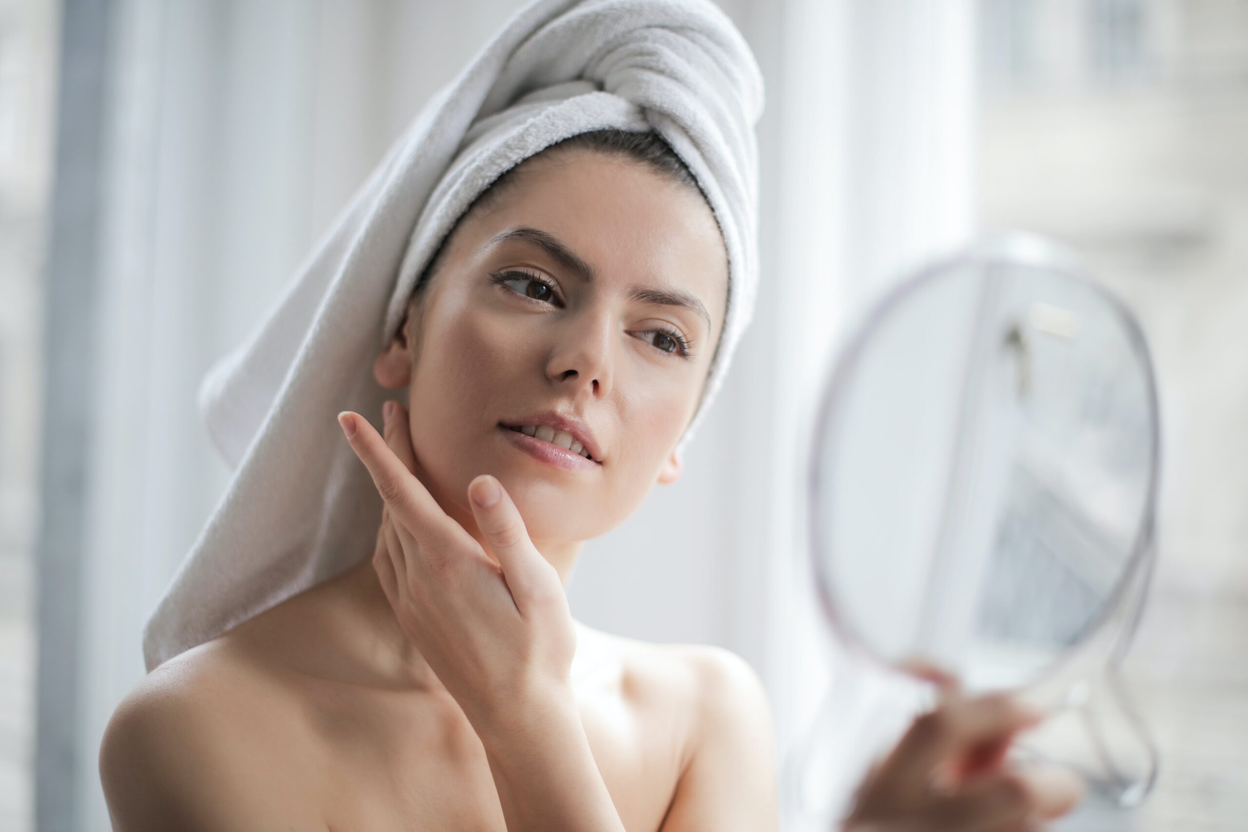 woman with towel on hair looking in handheld mirror touching face