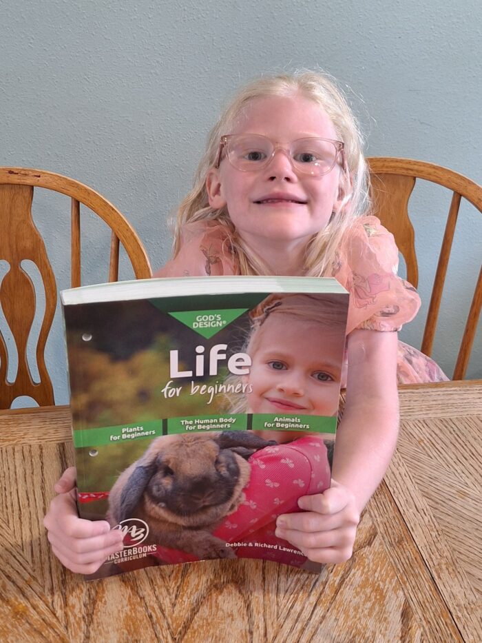 smiling girl wearing glasses holding up homeschool science book gods design life for beginners
