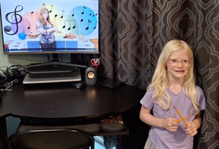 music teacher on computer monitor online class pointing to rhythm sticks on the table smiling girl holding pencils as rhythm sticks