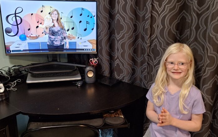 computer monitor with music teacher holding cat puppet and musical notes behind her smiling girl clapping her hands to the music
