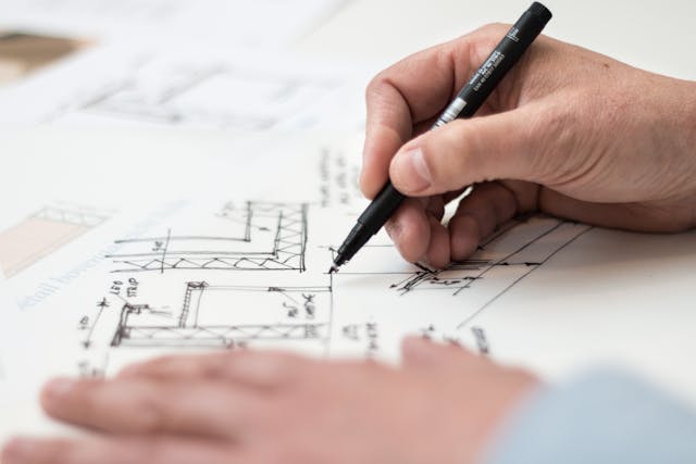 This article will explore the key factors to consider when trying to decide whether to buy, rent, or build your next home. home layout plan on blueprint with hand holding pen above it