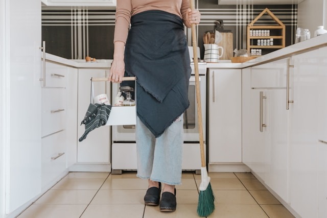 woman with apron and broom carrying cleaning caddy in kitchen