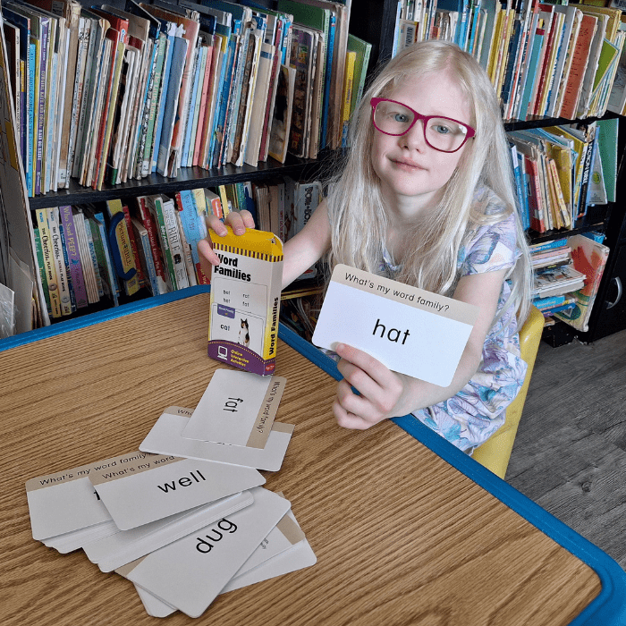girl wearing glasses holding a box of word families flash cards with word hat on a card and several cards on table