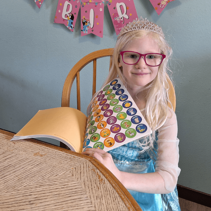 smiling girl wearing tiara and blue princess dress holding her workbook reward sticker page