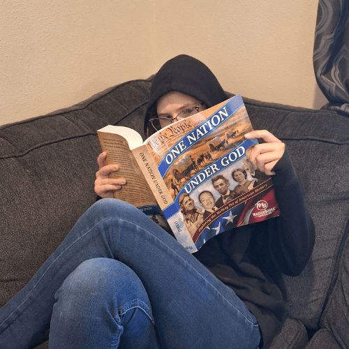 teen girl wearing hoodie and glasses sitting on couch with legs crossed reading one nation under god textbook