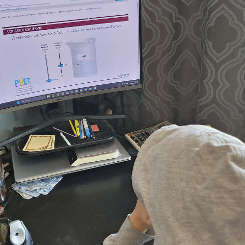 This is our product review of the Axiom Science High School Chemistry online course with 191 videos, 50 self-grading quizzes, and 20 Labs. student looking at computer monitor with saturated solutions and a beaker with liquid for her chemistry course